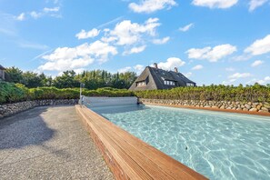 A clear swimming pool with a wooden deck, a stone wall, and a house with a thatched roof in the background. - Cozy 3-room vacation apartment, approximately 80 m², for 4 people. (Wenningstedt-Braderup (Sylt))