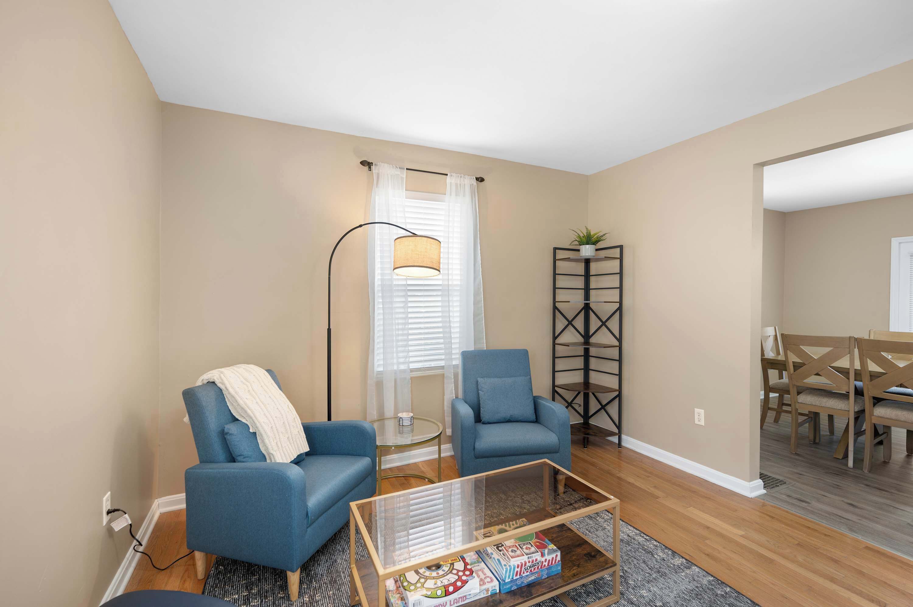 A living room with two blue armchairs, a glass coffee table, a floor lamp, and a dining area with wooden chairs.