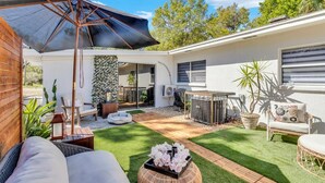 A modern outdoor patio with a covered seating area, a small garden, and a white building with large windows. - Backyard Zen  Retreat with Modern Vibe (Tampa)