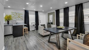 A modern living room with a dining table, a sofa, a kitchen area, and a large window with curtains. - Backyard Zen  Retreat with Modern Vibe (Tampa)