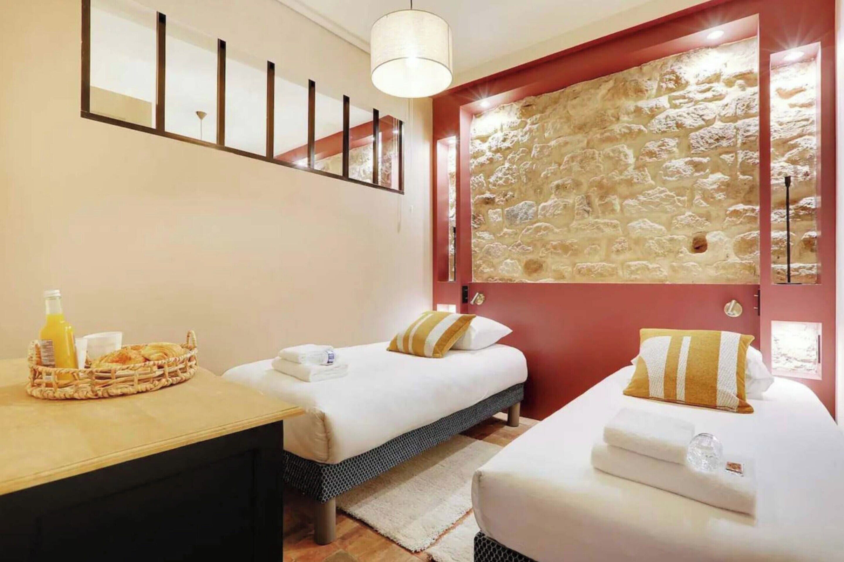 A hotel room with two beds, a stone wall, a wooden desk with breakfast items, and a mirror above the bed.