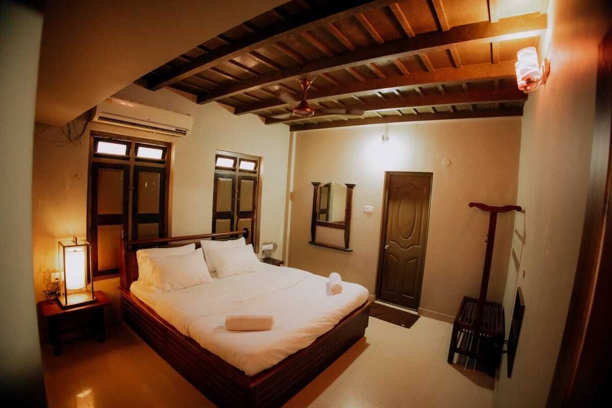 A bedroom with a wooden ceiling, a bed with white linens, a nightstand with a lamp, and a door with a mirror.