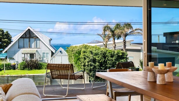Een modern terras buiten met een houten tafel en stoelen, met uitzicht op de kustlijn met een huis en palmbomen.