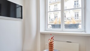 A room with a large window, a radiator, and a vase with flowers on a small table. - Charming Studio - Louvre & Palais Royal (Paris)