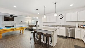 Une cuisine moderne avec des meubles blancs, un îlot central doté de tabourets de bar, une table de billard intégrée et des luminaires suspendus.