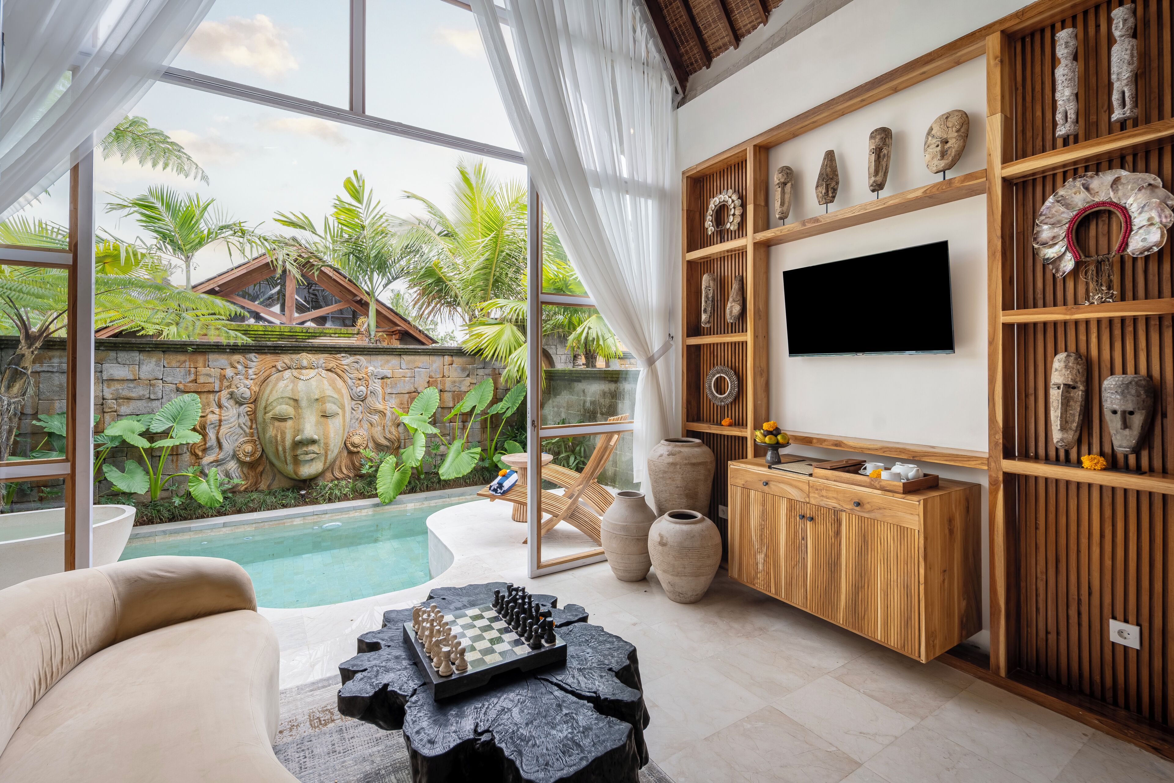 A modern living room with a pool view, wooden shelves with decorative items, and a flat-screen TV.