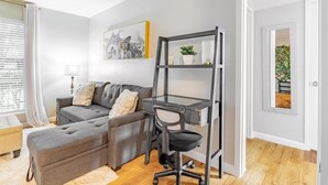 Ein modernes Wohnzimmer mit einem grauen Sofa, einem Schreibtisch mit Glastischplatte und einem Sessel. An der Wand hängt ein Gemälde und an der Tür ein Spiegel.