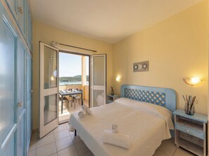 A bedroom with a bed, a nightstand, a desk with chairs, and a view of the sea through an open door. - Relax in Marinella With sea View (Golfo Aranci)