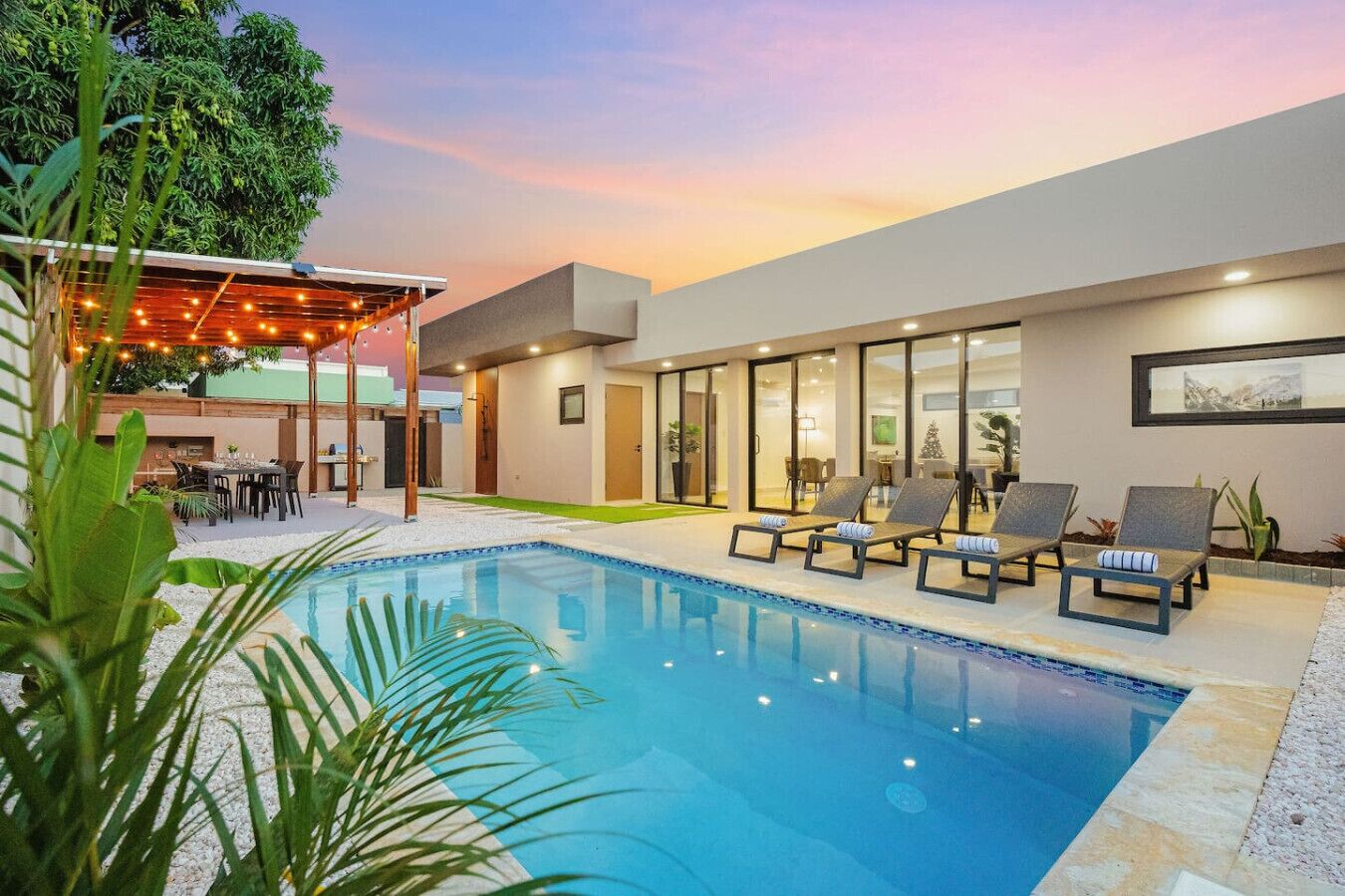 A modern pool area with lounge chairs, a pergola with string lights, and a view of the property.
