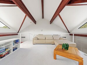 A spacious attic room with a white ceiling, wooden beams, a beige sofa, a wooden coffee table, and a bookshelf with books. - Sailors Hill Retreat—Bushland Living by the Coast (Naturaliste)