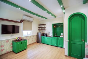 A kitchen with green cabinets, a refrigerator, a microwave, and a sink. There is a window with blinds and a flat-screen TV mounted on the wall. - Casa Inali (New Delhi)