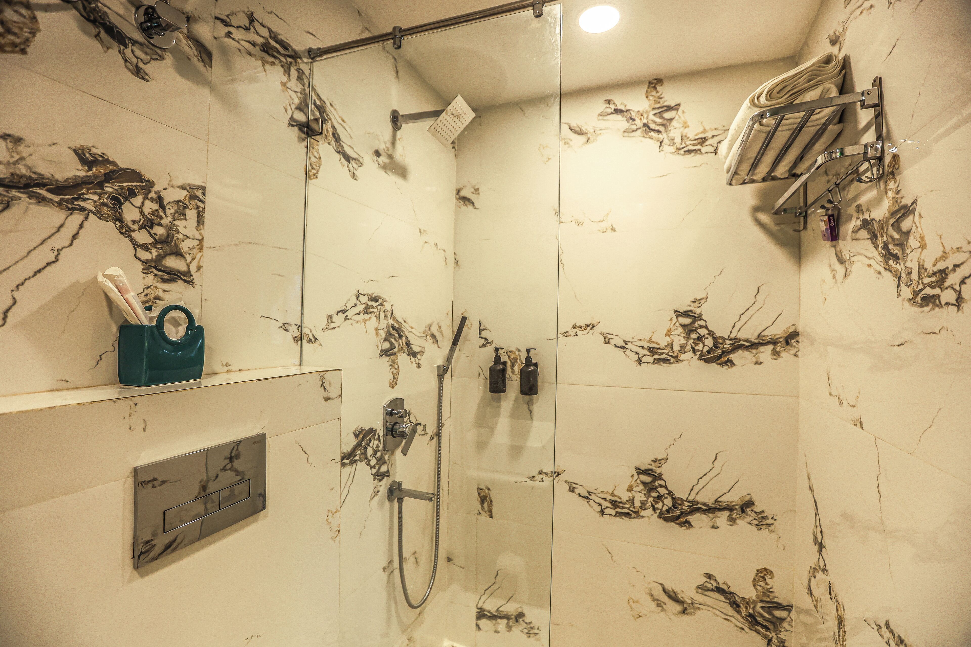 A bathroom with a glass shower enclosure, marble-patterned walls, a towel rack, and a soap dispenser.