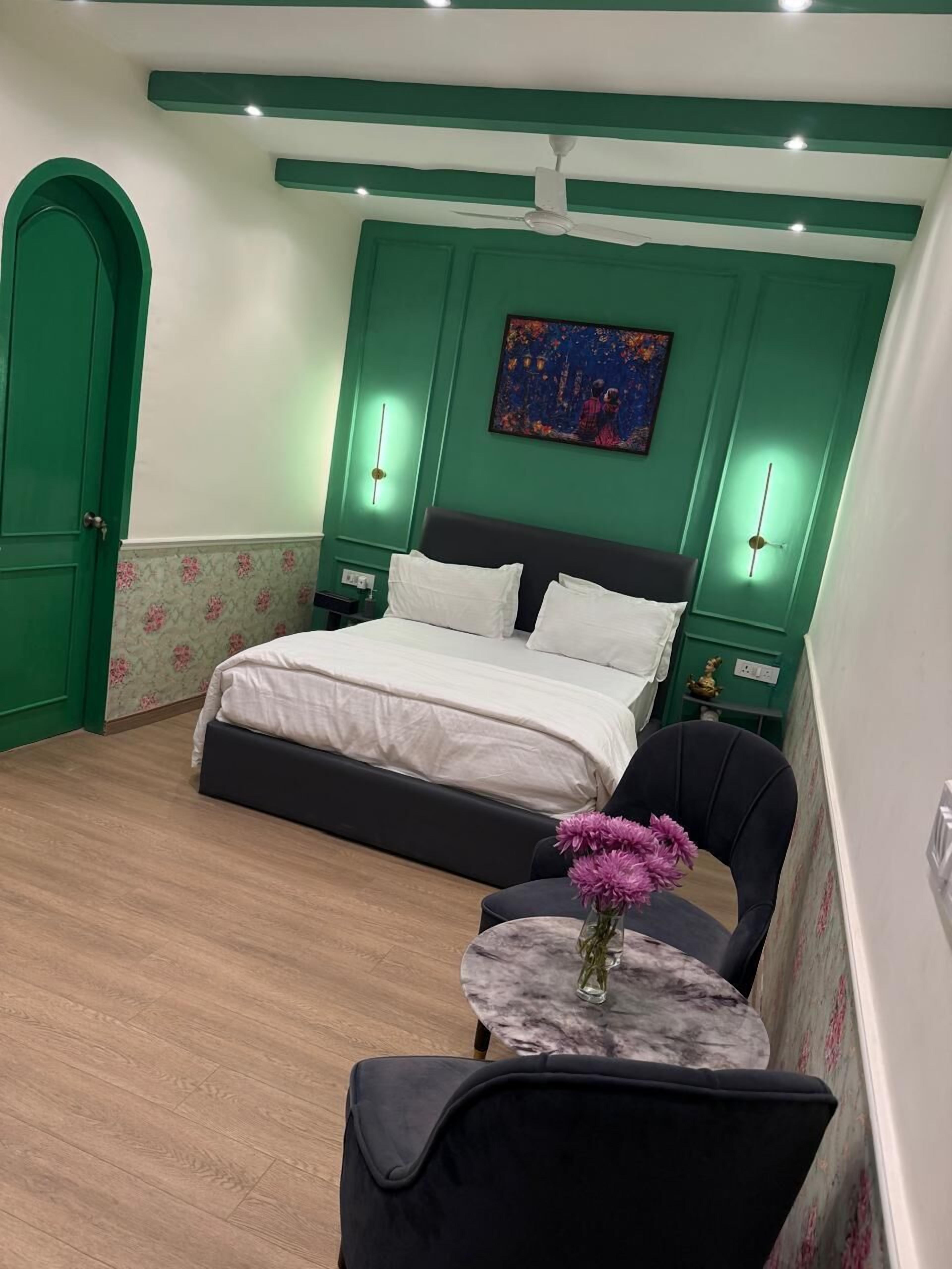 Een slaapkamer met een bed, een stoel, een klein tafeltje met bloemen en een schilderij aan de muur.