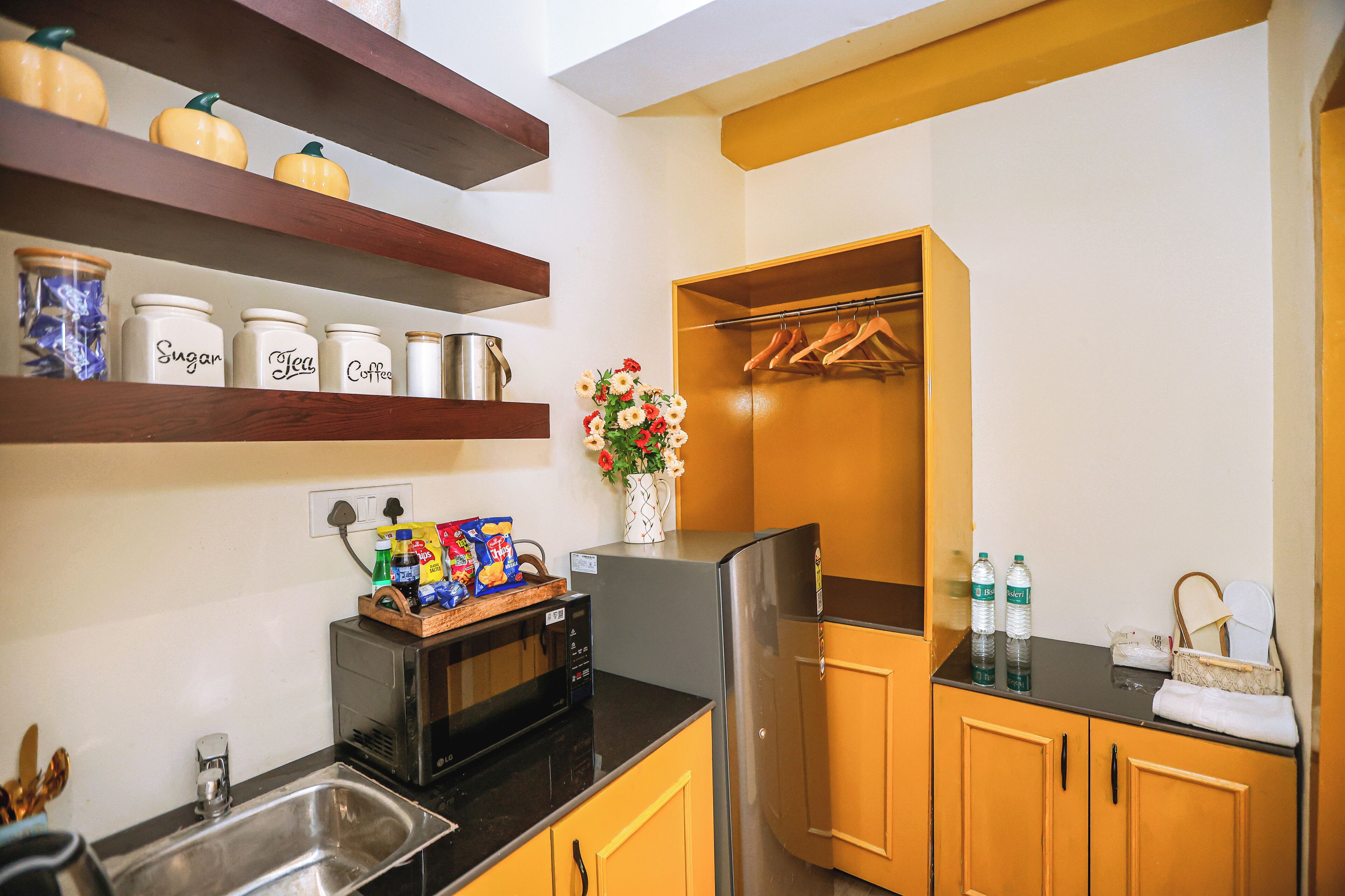 A compact kitchen with wooden cabinets, a stainless steel refrigerator, and a microwave. There are shelves with jars labeled "Sugar," "Tea," and "Coffee." A small wooden table holds snacks and a vase with flowers.