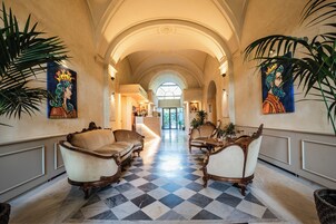 Um hall de hotel com piso xadrez, móveis elaborados e grandes painéis nas paredes.