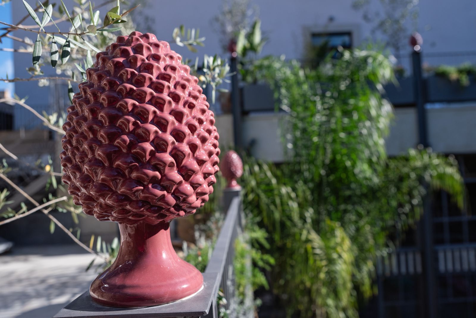Eine rote, strukturierte Skulptur auf einem Metallfuß, mit einem Gebäude und Grünpflanzen im Hintergrund.