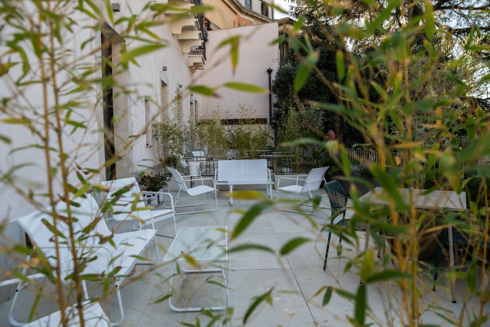 Pátio externo com móveis de metal branco, incluindo cadeiras e uma mesa, rodeado por vegetação e um edifício ao fundo.