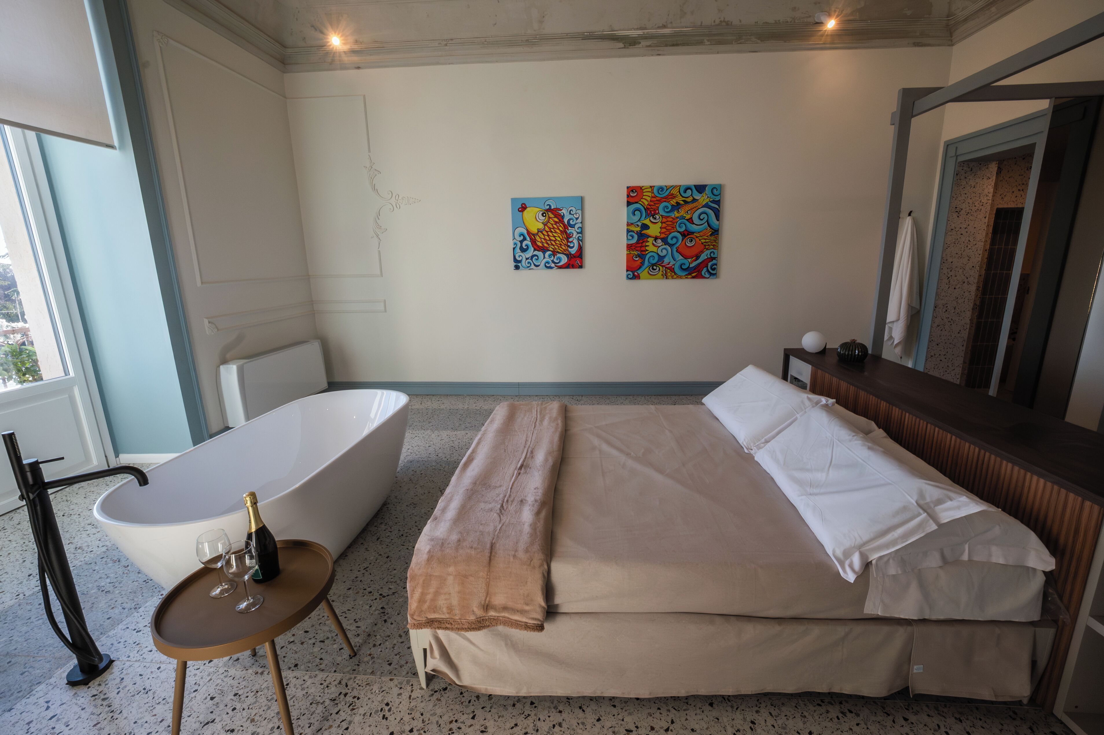 Een moderne badkamer met een vrijstaand bad, een bed met beige beddengoed en twee kleurrijke schilderijen aan de muur.