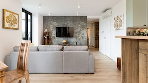 A modern living room with a grey sofa, wooden dining table, and a TV mounted on a textured wall. - Araliya (Kraków)