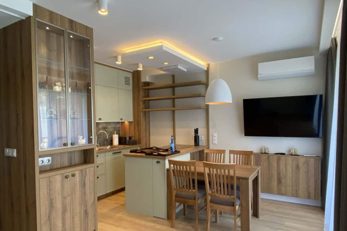 A modern kitchen with wooden cabinets, a dining area with a table and chairs, and a flat-screen TV mounted on the wall.