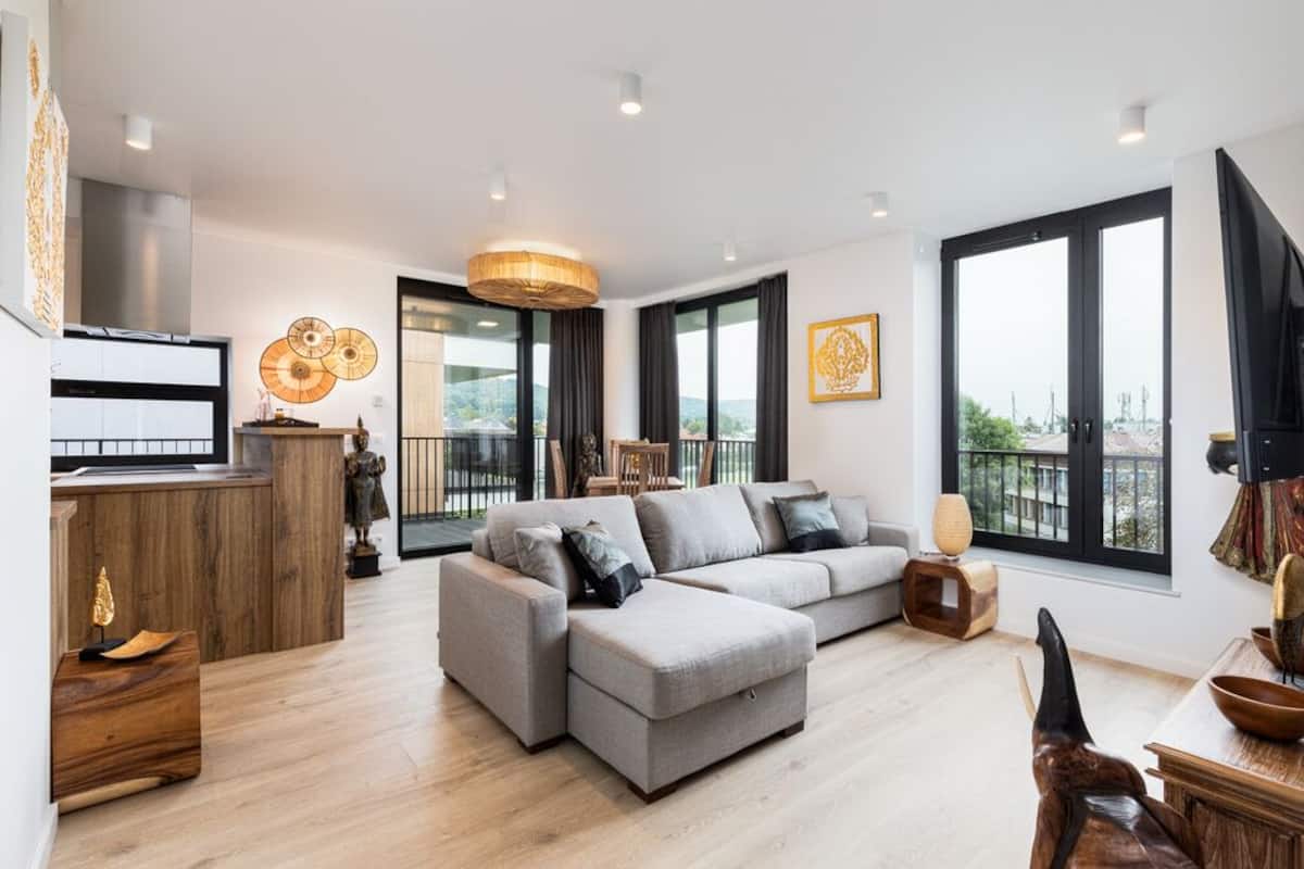 A modern living room with a grey sectional sofa, a wooden coffee table, and a dining area with a wooden table and chairs. There is a large window providing a view of the outdoors.