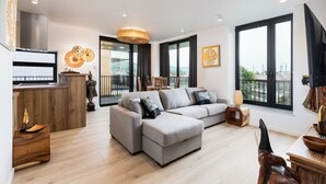 A modern living room with a grey sectional sofa, a wooden coffee table, and a dining area with a wooden table and chairs. There is a large window providing a view of the outdoors. - Araliya (Kraków)