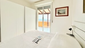 Et soveværelse med en seng, en dør der fører til en balkon, et billede i ramme på væggen og et lille bord med en lampe.
