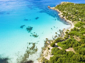 Vista aérea de una zona costera con aguas turquesas cristalinas, playa de arena y vegetación verde exuberante.