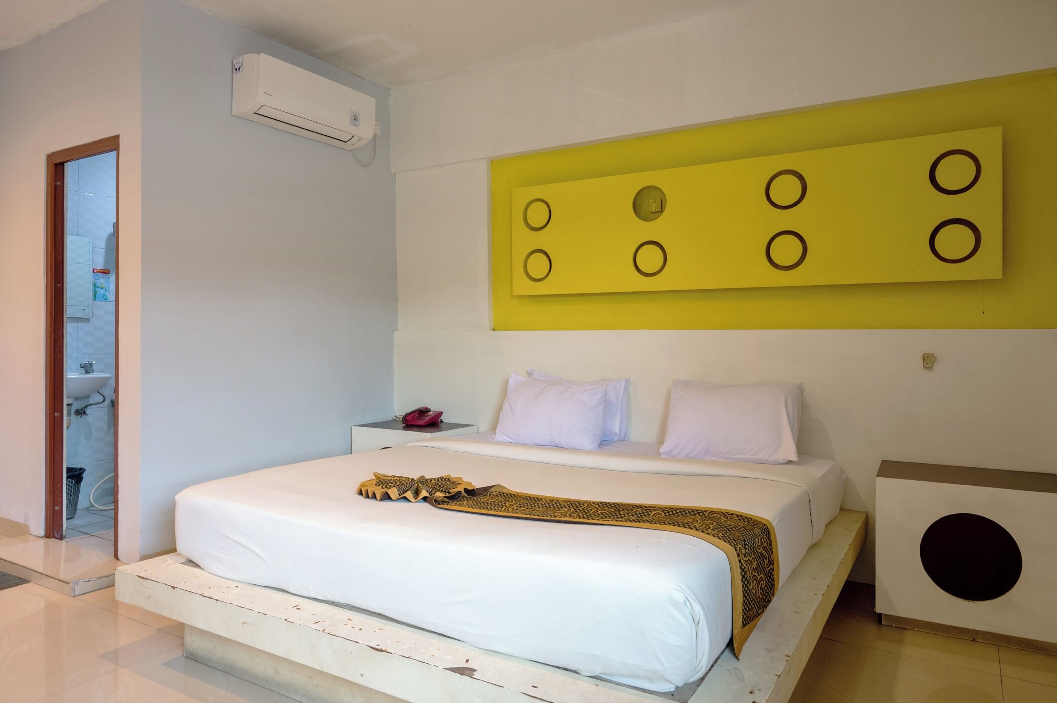 A bedroom with a bed, a bedside table, an air conditioning unit, and a unique yellow wall panel.