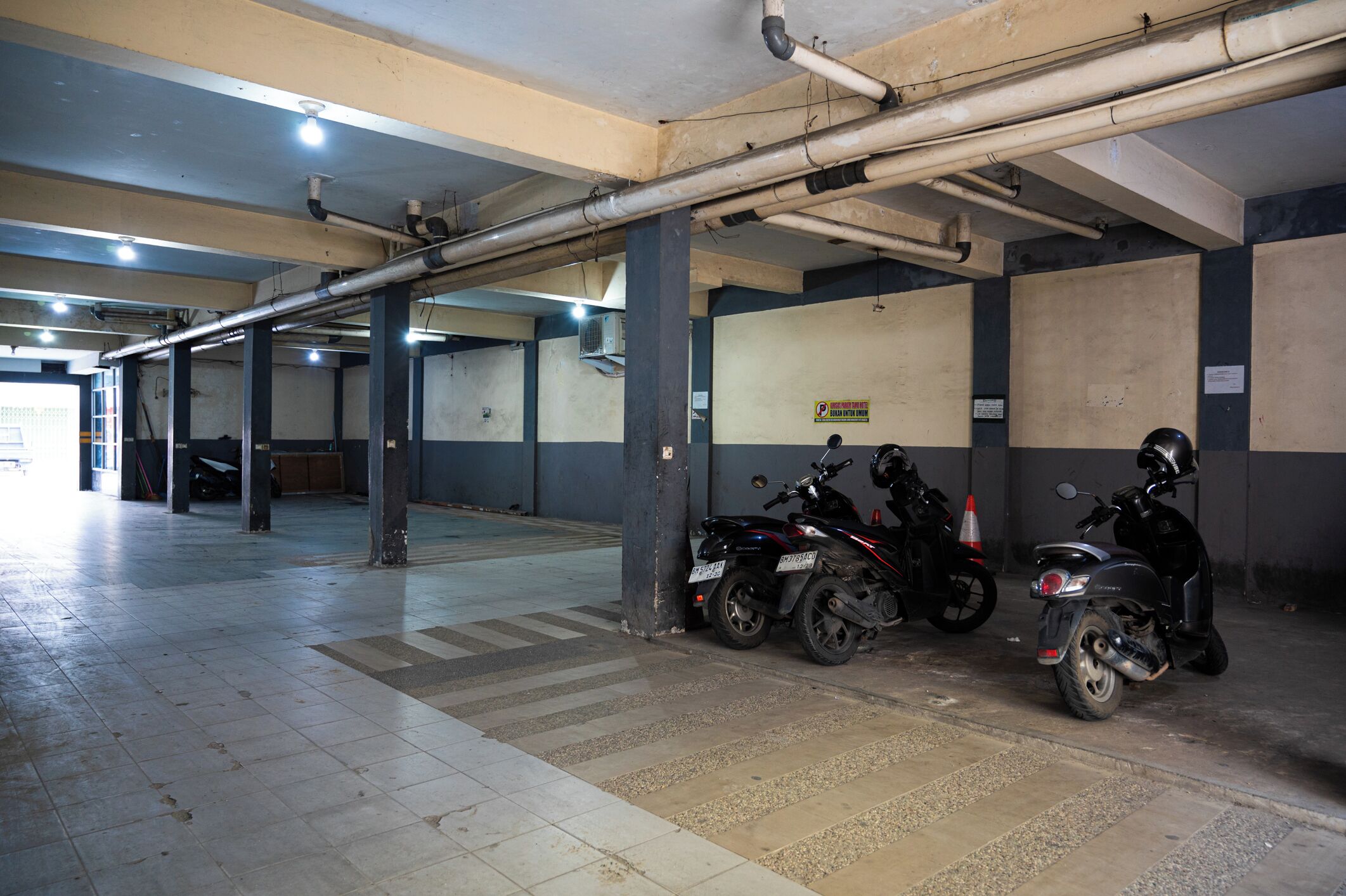 Un estacionamiento interior con pisos y paredes de concreto, que muestra dos scooters aparcados y una unidad de aire acondicionado visible.