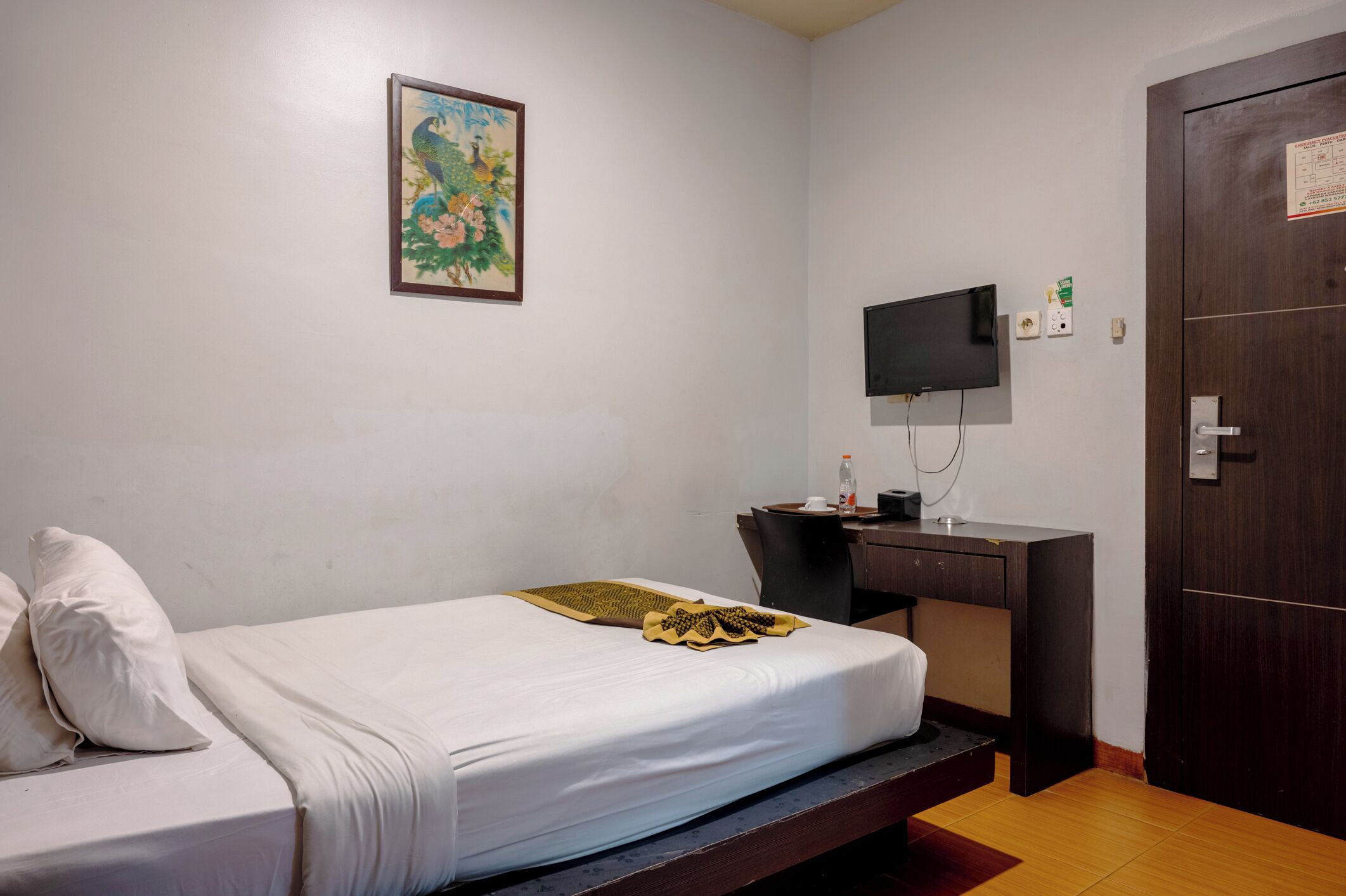 A single bed with a white mattress and pillows, a desk with a chair, a television mounted on the wall, and a wooden door.