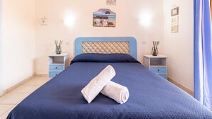 Un lit bien fait, avec une tête de lit bleue et une serviette enroulée posée dessus. Il y a deux tables de chevet ornées d’objets décoratifs et un petit tableau accroché au mur au-dessus du lit.