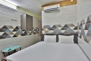 A hotel room with a bed, a small table with a remote control, a wall-mounted air conditioner, and a patterned wall.