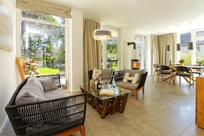 A spacious living area with a glass coffee table, comfortable seating, a fireplace, and large windows offering a view of the outdoors. - Exclusive 5-star vacation home with a sauna, hot tub, and garden in Karlshagen (Karlshagen)