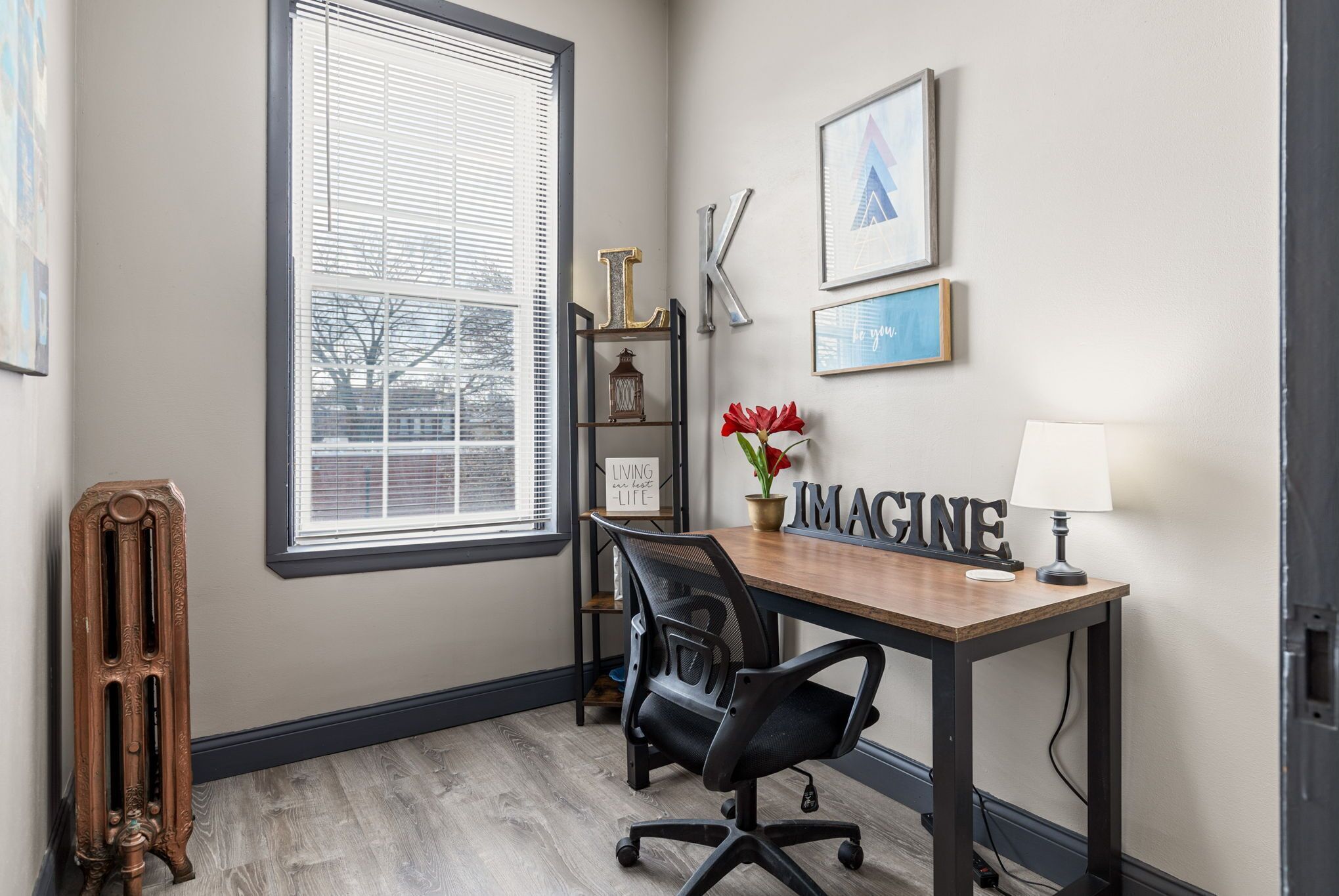 A dedicated workspace offers a desk, office chair, lamp, artwork, radiator, and abundant natural light.