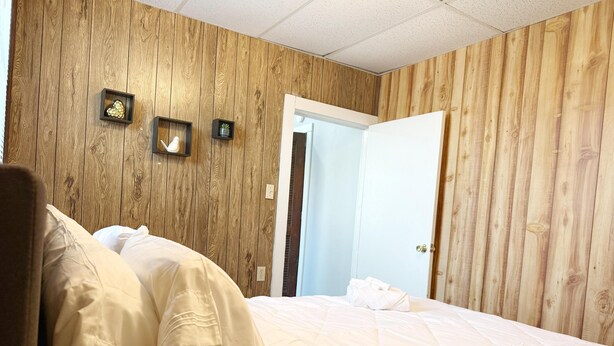 Een slaapkamer met een houten wandpaneel, een bed met wit beddengoed en een klein plankje met decoratieve objecten.