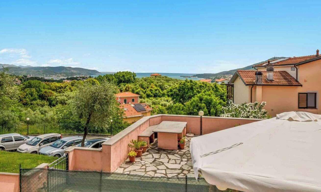 Eine Dachterrasse mit Steinboden, einem Tisch und Stühlen mit Blick auf eine Küstenlandschaft, Bäume und geparkte Autos.