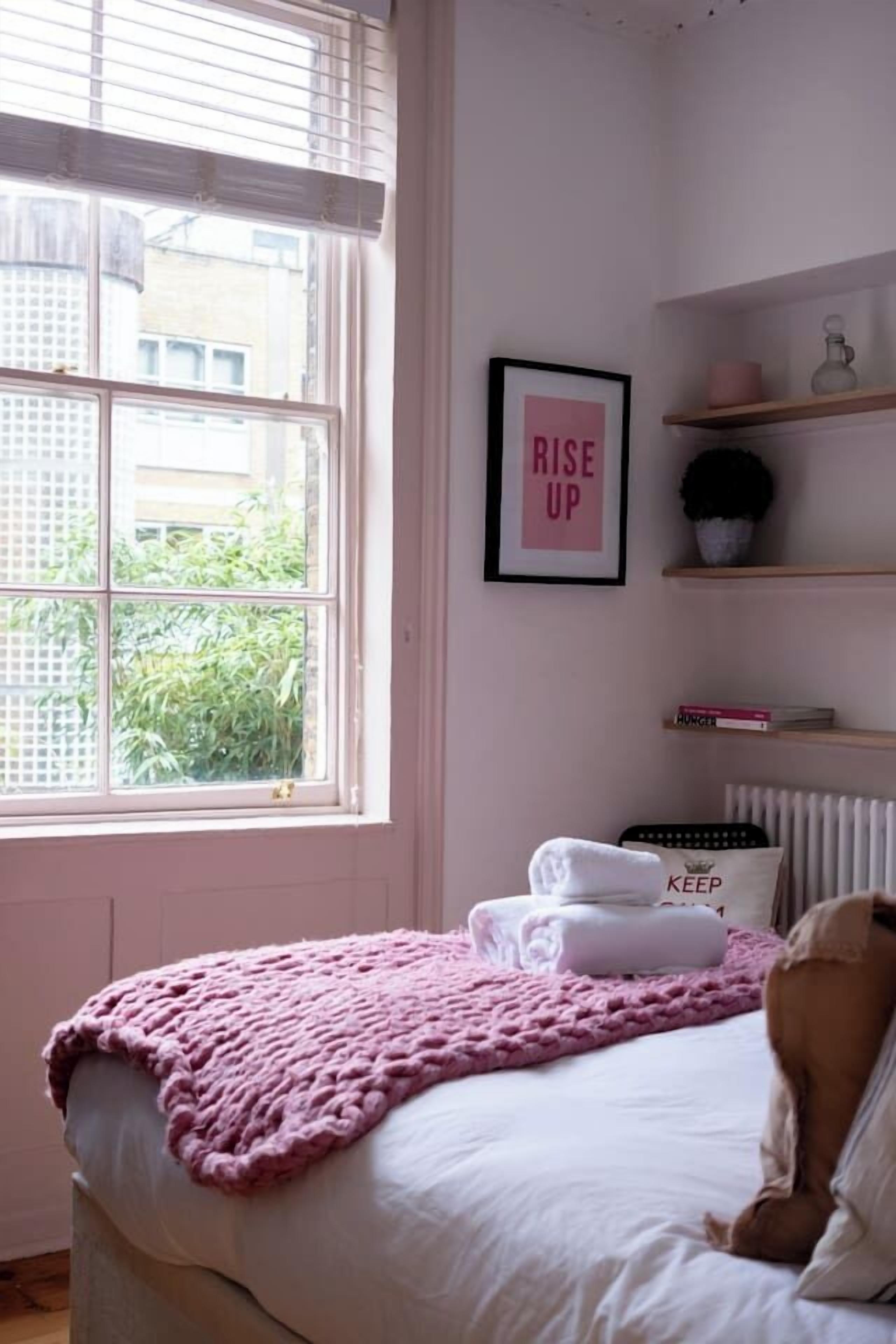 A bedroom with a bed, a window, a radiator, a shelf with books, and a framed motivational poster.