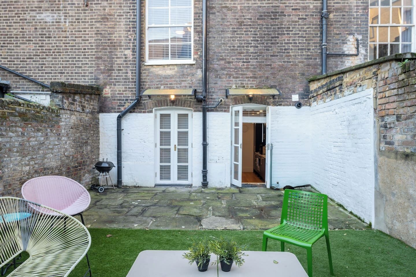 A courtyard with a brick wall, a white door, a green chair, and a table with plants.