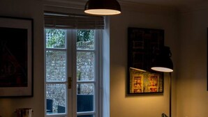 Ein Essbereich mit Tisch, Stühlen und einer Pendelleuchte. Ein Fenster mit Jalousien, eine Couch und ein Gemälde an der Wand sind ebenfalls vorhanden.
