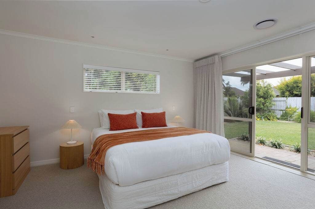 A bedroom with a large bed, a bedside table with a lamp, a window with blinds, and a sliding glass door leading to a garden.