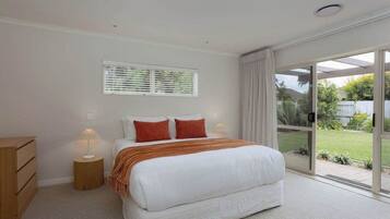 A bedroom with a large bed, a bedside table with a lamp, a window with blinds, and a sliding glass door leading to a garden.