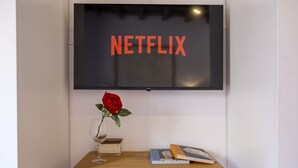 En platt-TV är monterad på en vägg och ordet ”NETFLIX” visas på skärmen. Under TV:n står ett träbord med en vas av glas som innehåller en röd ros, ett vinglas och några böcker.