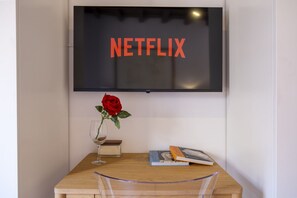 Ein wandmontierter Flachbildfernseher zeigt 'NETFLIX' an. Darunter ein Holztisch mit einer Glaskaraffe, in der eine rote Rose steht, ein Weinglas und einigen Büchern.