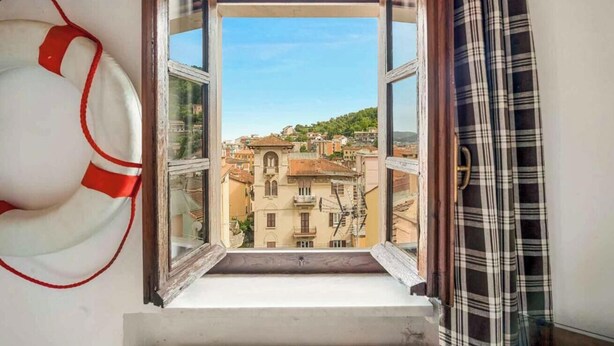A window with a view of a cityscape, a life preserver hanging on the wall, and a plaid curtain partially drawn. - Welcome to La Goletta, your holiday home on the third and top floor of a quiet building in La Spezia. This cozy two-room apartment is perfect for families, couples or small groups of friends.🛏 Sleeps up to 4 people with a double sofa bed in the living (La Spezia)