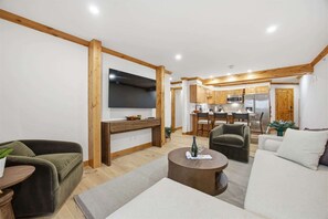 Ein modernes Wohnzimmer mit Flachbildfernseher, einer Couch, einem Couchtisch und einer Küche im Hintergrund.