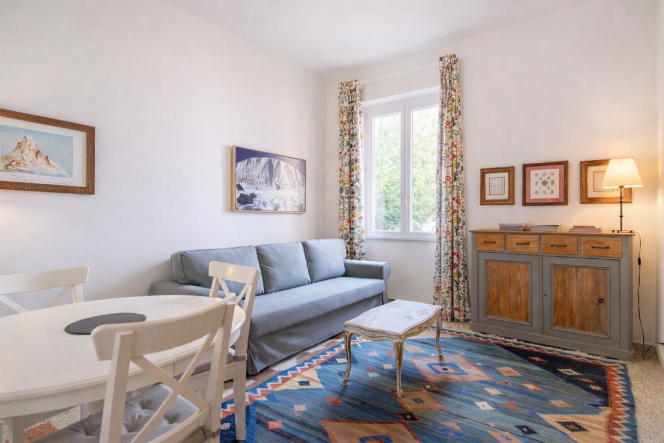 Ein Wohnzimmer mit einer Couch, einem Esstisch mit Stühlen, einem gemusterten Teppich, einer Kommode mit Schubladen und gerahmten Bildern an der Wand.