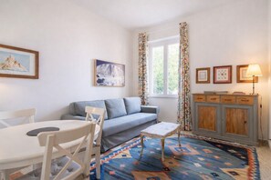 Ein Wohnzimmer mit einer Couch, einem Esstisch mit Stühlen, einem gemusterten Teppich, einer Kommode mit Schubladen und gerahmten Bildern an der Wand.