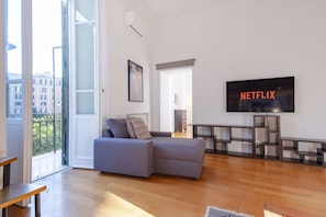 A modern living room with a grey sectional sofa, a flat-screen TV mounted on the wall, and a wooden floor. - Elegantis.it Ten windows on Trastevere (Roma)