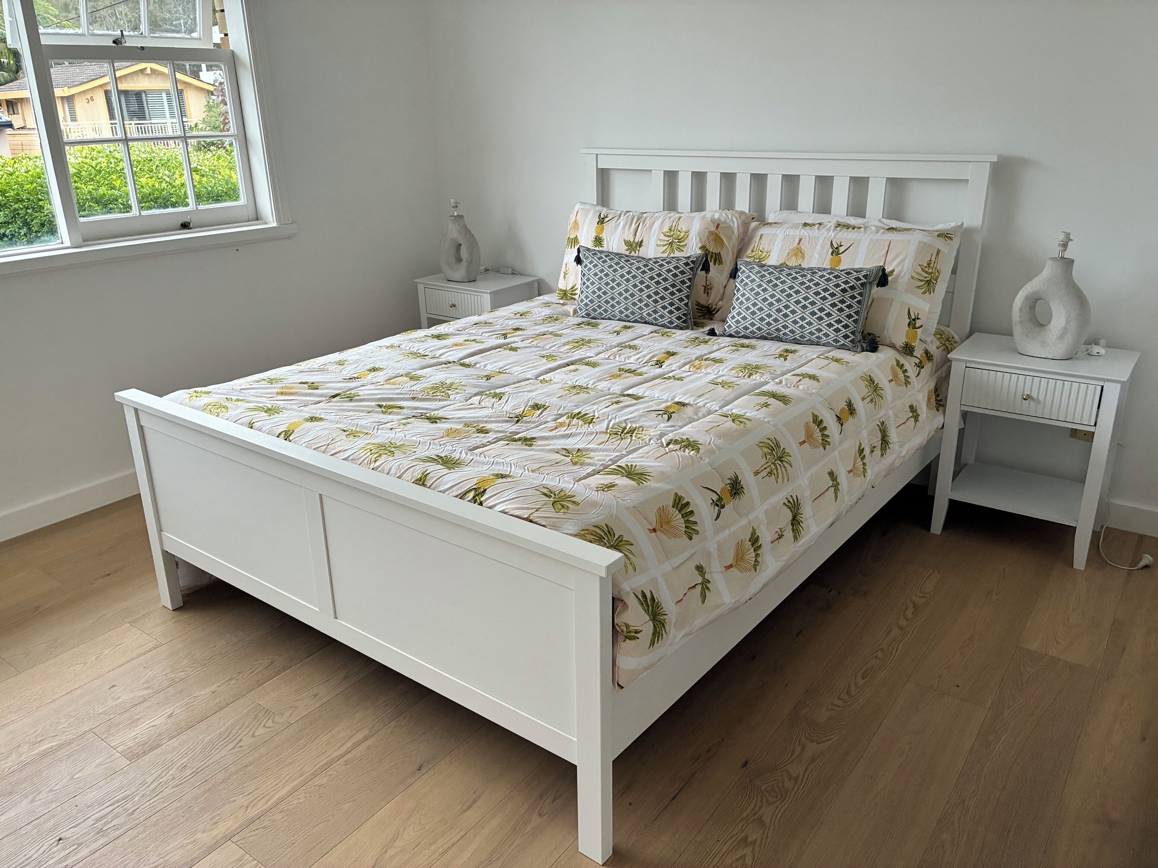 A white bed with a leaf-patterned bedspread, two matching pillows, a nightstand with a vase, and a window with a view of greenery.